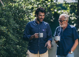 Younger and older man in blue shirts holding coffee cups outside chatting