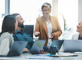 Woman in tan jacket presenting to co-workers around a table with laptops